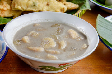 Kolak pisang, traditional Indonesian dessert made of bananas cooked in coconut milk and palm sugar syrup, served  in a white bowl. Setting appears to be a wooden table. 