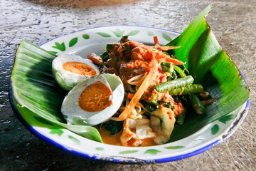 Indonesian vegetable salad with peanut sauce or pecel with  salted egg on vintage enamel plate  lined with banana leaves, places on ethnic fabric