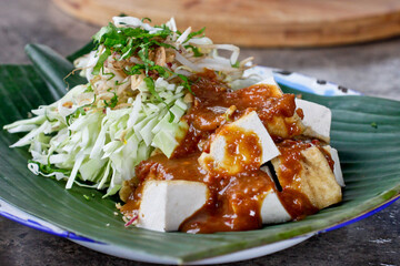Indonesian tahu campur. Tofu pieces topped with a rich brown sauce are artfully arranged on a banana leaf. Accompanying the tofu are fresh, thinly sliced cabbage and bean sprouts