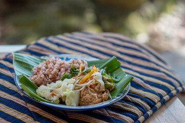 Indonesian vegetable salad with peanut sauce or pecel with brown rice on vintage enamel plate  lined with banana leaves, places on ethnic fabric