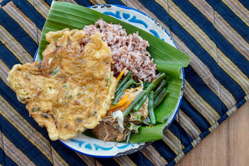 Indonesian vegetable salad with peanut sauce or pecel with brown rice and omelet on vintage enamel plate  lined with banana leaves, places on ethnic fabric