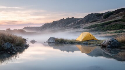 Serene camping scene shows a yellow tent reflected in a misty mountain lake at sunrise providing an outdoor adventure