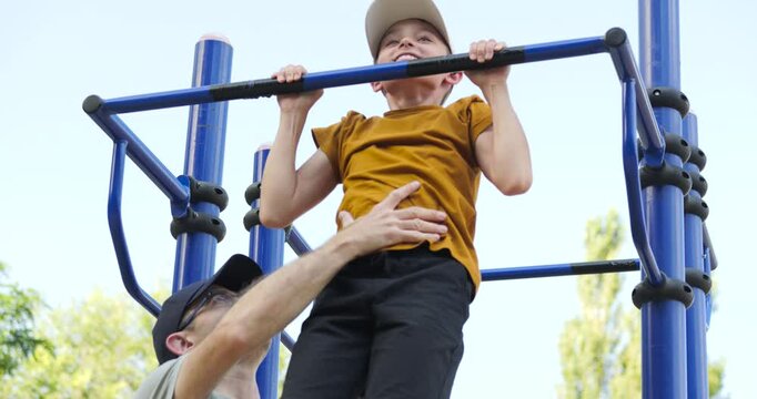 Portrait child doing pull-ups on horizontal bar on sports ground. child learning to do pull-ups on horizontal bar. active child doing sports with his father outdoors. Family sports on sports ground.