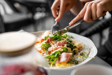 Close-up of a person eating a fresh salad with ham, greens, tomatoes, and cheese at a restaurant table, using fork and knife. Concept of healthy and appetizing meal.