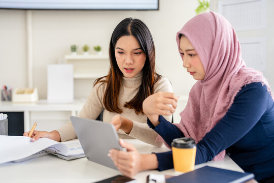 Business woman in hijab and colleague collaboratively review content on a tablet in a bright office space.