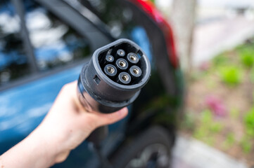 Person grips electric vehicle charger with car partially visible in background, parked in...