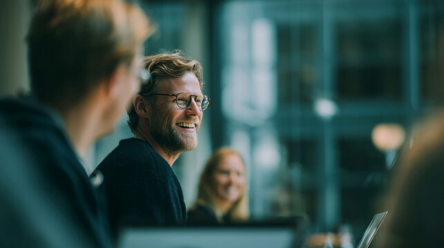 Laughing colleagues with laptop and tablet in office, happy teamwork and collaboration in casual meeting with soft natural daylight