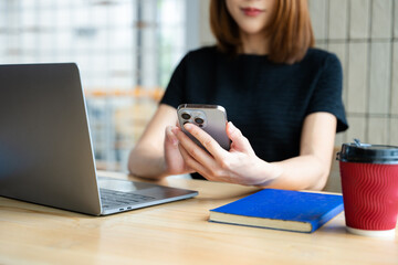 Asian businesswoman using smartphone and laptop in a modern workspace by a window, showcasing...