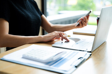 Asian businesswoman using smartphone and laptop in a modern workspace by a window, showcasing...