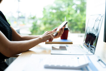 Asian businesswoman using smartphone and laptop in a modern workspace by a window, showcasing...