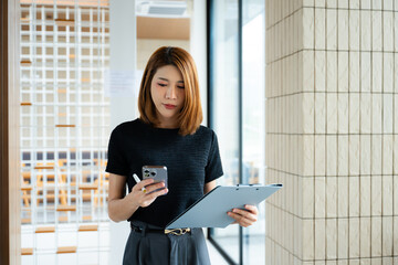 Asian businesswoman taps smartphone in a modern office workspace, highlighting digital...