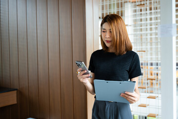 Asian businesswoman taps smartphone in a modern office workspace, highlighting digital...