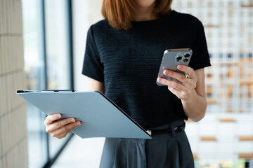 Asian businesswoman using smartphone and laptop in a modern workspace by a window, showcasing...