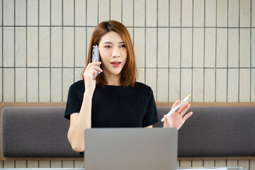 Asian businesswoman talking on smartphone while holding a pen and reviewing a laptop in a modern...