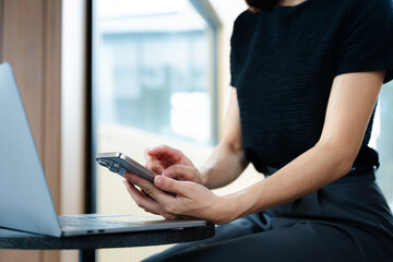 Asian businesswoman using smartphone and laptop in a modern workspace by a window, showcasing...