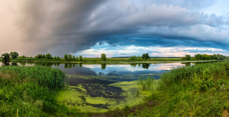 Panoramic view of the river from the shore with dark storm clouds in the sky. Scenic non-urban landscape of nature. Deciduous trees and green grass