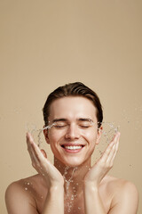 Young man washing face with water splash on beige background, copy space