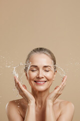 Mature senior woman washing face with water splash on beige background, copy space