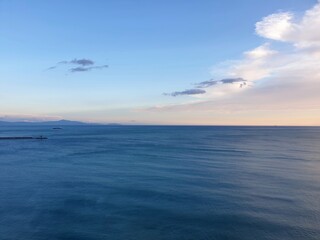 A calm ocean stretches to the horizon. A breakwater is to one side, with a boat framed by distant hills. The water is dark blue, and the clouds have the colours of sunset.