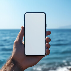 Image of male hand holding white mobile phone with blank screen sitting on seashore against blue sky background.