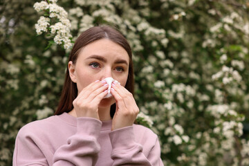 Woman with paper tissue suffering from seasonal pollen allergy near blossoming shrub on spring day,...