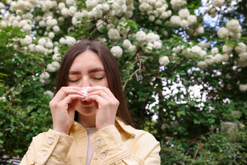 Woman with paper tissue suffering from seasonal pollen allergy near blossoming shrub on spring day,...