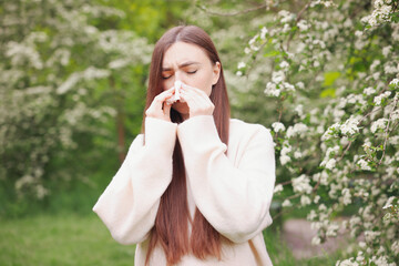 Woman with paper tissue suffering from seasonal pollen allergy near blossoming shrub on spring day