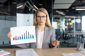 A frustrated businesswoman in a gray suit holds a chart with a negative trend, expressing concern...