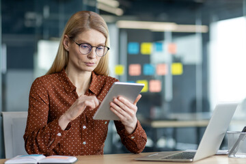 A businesswoman is working at her desk with a tablet and laptop in a modern office setting, wearing...