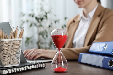 Deadline. Woman with laptop at wooden table in office, focus on hourglass