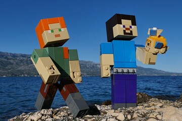 Naklejka premium LEGO Minecraft Steve holding LEGO small and large Pufferfishes aquatic mobs, Alex standing next to him. Location rocky Croatian beach, bay water and Velebit mountain in background. Blue summer skies. 