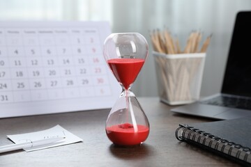 Deadline. Hourglass with flowing sand, stationery, calendar and laptop on wooden table indoors