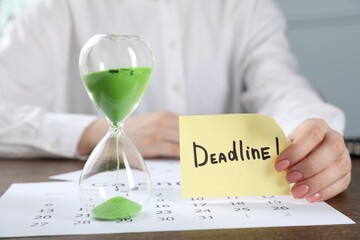 Woman holding sticky note with word Deadline at wooden table in office, closeup