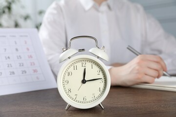 Deadline. Woman writing notes at wooden table in office, focus on alarm clock