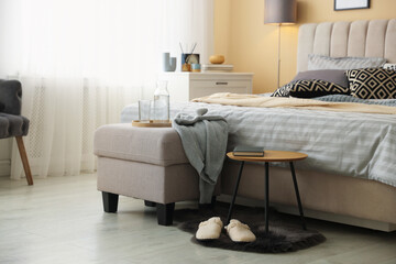 Comfortable bed with soft linens, slippers, pouf and side table in room. Home textile