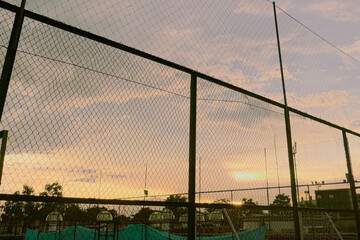 A chain-link fence in silhouette against a beautiful sunset sky with clouds and warm light.