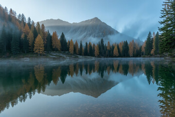 Fototapeta premium Mountains and trees reflect in a serene lake under a misty sky