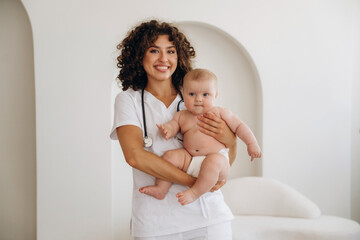 In coat, with stethoscope. Holding kid in hands. Female doctor is with newborn baby indoors