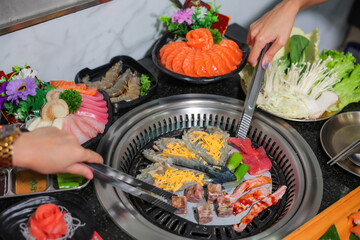 A woman's hand holding meat tongs with Full set of beef and pork ready for grill on stove serve with vegetable, soup and rice and side dish, barbecue style.	
