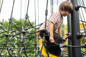 A child boy is climbing up an alpine grid in a park on a playground on a hot summer day. Kids playground in a public park, entertainment and recreation for children, mountaineering training.
