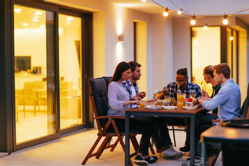 A group of young diverse people having dinner on the terrace of a modern house in the evening. Fun...