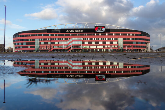 Alkmaar, Netherlands. 11-26-2024. The AFAS Stadium, home of AZ Alkmaar, reflects beautifully in a puddle on a cloudy day, showcasing its striking red and black design under dramatic skies.