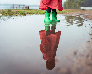 A playful child in green boots and a red raincoat splashes in a puddle reflecting their image
