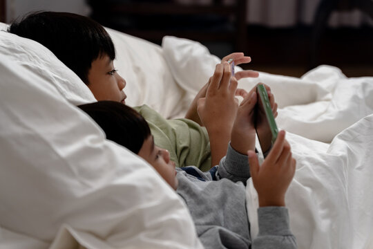 Two children are laying on a bed and playing with their cell phones