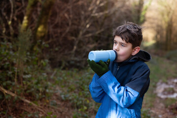 A young boy drinks from a blue water bottle while standing in a green forest, he is dressed warmly...