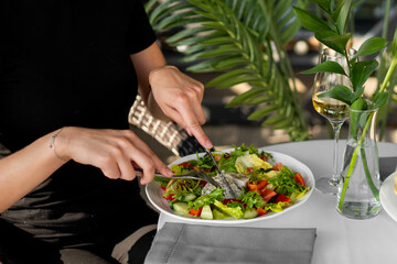 Person cutting fresh vegetable salad at a restaurant table with white wine and decorative greenery. Casual dining atmosphere, healthy lifestyle and gourmet food concept.