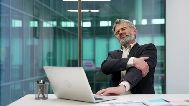 Mature bearded businessman suffering from elbow pain sitting in a business office. Upset male in suit is massaging a joint, holding a sore spot with his hand. He has a joint injury or ligament sprain