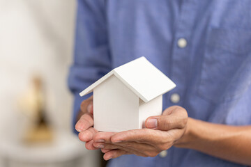 Person holding a small white wooden house with both hands, symbolizing home ownership, real estate,...