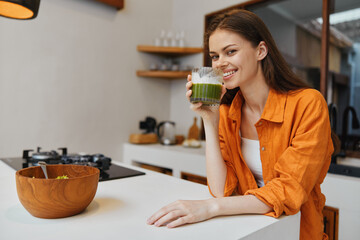 Young woman enjoying a healthy green smoothie in a modern kitchen, wearing an orange shirt and...