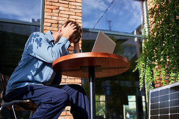 Man works on laptop at outdoor round table, holding head in frustration. Solar panel nearby,...